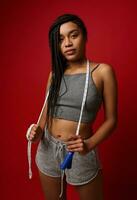 Confident African woman with stylish dreadlocks and fit body posing against red background with jumping rope. Cardio workout, weight loss and fitness training concept with copy ad space photo