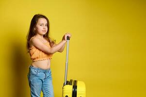 Beautiful little traveler girl with long hair, dressed in blue jeans and orange top, with suitcase, yellow background. photo
