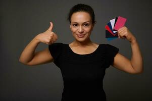 Pretty woman in black smiles with beautiful smile and poses against gray background with many discount or credit cards in her hands showing thumb up looking at camera. Shopping concept at Black Friday photo