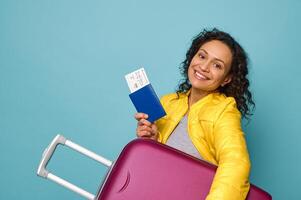 Charming woman traveler in bright yellow jacket, smiling cheerful toothy smile, holding suitcase under arms, showing her passport and ticket with boarding pass to camera. Blue background copy space photo