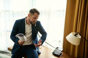 Young handsome Caucasian businessman, arguing while talking on landline phone, holding documents in his hand and gesturing with hand during negotiations by phone, sitting on the desk corner in hotel photo