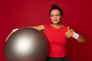 Portrait of Hispanic pretty fit woman holding a fit ball and showing a thumb up looking at camera. Copy free space for text on red colored background. photo