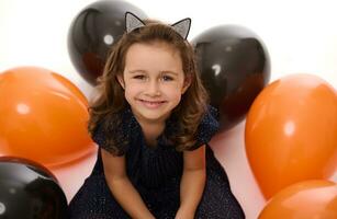 Close-up portrait of a cute little girl smiling with beautiful toothy smile looking at camera on the white background with lying down inflated black and orange colored balloons. Halloween concept photo