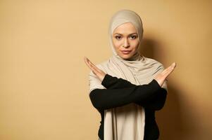 Muslim woman crosses her arms to show denial, beige background with copy space photo