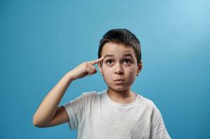 Pensive boy pointing with forefinger to temple while standing on blue background with copy space photo