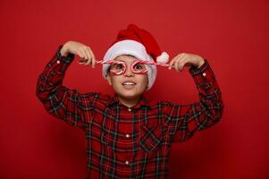 Adorable boy in Santa Claus hat and plaid shirt looks at the camera through sweet lollipops, holding them imitating eyeglasses, posing on a colored background with copy space for Christmas advertising photo