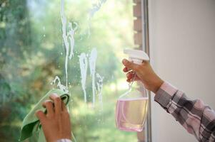 de cerca de el manos de un mujer limpieza el ventana, quitando manchas por pulverización limpieza productos y limpiando con un esponja. primavera casa limpieza concepto con Copiar anuncio espacio foto