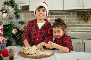 Beautiful kids looking at camera while cooking together in the home kitchen during Christmas holidays. Charming preadolescent boy in Santa hat and his cute little sister kneading dough for bread photo