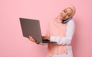 Concept of a busy Arab muslim business woman, freelancer in hijab, talking on a mobile phone while working on a laptop at the same time. Isolated on pink background with copy space photo