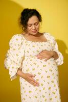 Pregnant pretty woman in sundress, gently caresses her belly, experiencing happy moments of her carefree happy pregnancy photo