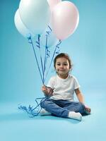 linda pequeño niña sentado en azul antecedentes con suave sombra y posando a cámara con globos en el mano. infancia y niño proteccion día concepto. Copiar espacio foto