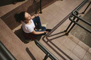 alto ángulo ver de un mujer sentado en patineta con auriculares y teléfono inteligente en su mano y tomando Dom baños al aire libre foto