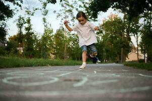 longitud total retrato de un adorable pequeño niña jugando rayuela en el suelo de un ciudad parque en un hermosa calentar verano día a puesta de sol. calle para niños juegos en clásicos foto