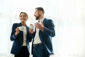 Two business people, partners, colleagues having a conversation at coffee break near the window in the meeting room. Attractive middle aged woman and handsome young man enjoying coffee break together. photo