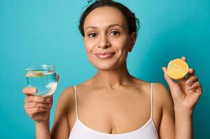 hermosa africano americano mujer participación un medio de Fresco jugoso amarillo limón y un vaso con agua en su manos, sonriente lindo mirando a cámara aislado terminado azul antecedentes con Copiar espacio para anuncios foto