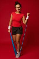 Attractive athletic fit woman doing body weight workout using elastic strap pulling with arms for shoulder training against red background with copy ad space photo