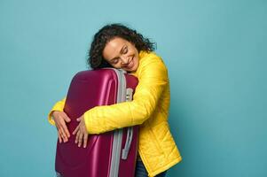 Cheerful African American woman in bright yellow jacket hugging her suitcase, smiling with cheerful toothy smile looking at camera, isolated on blue background with copy ad space photo