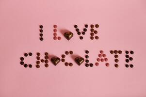 Top view of I LOVE CHOCOLATE lettering laid out from multi colored chocolate pills and candies on pink background with copy space photo