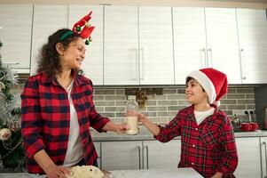 hermoso preadolescente chico en Papa Noel sombrero trayendo un botella con Leche a su amoroso madre Cocinando masa para Navidad galletas en el hogar cocina. hijo Ayudar su mamá en horneando foto