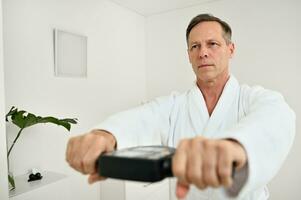 Portrait of a handsome middle aged male patient in white terry bathrobe weighing himself using a medical equipment - smart scale to determine biological age and vitality at a spa wellness center photo