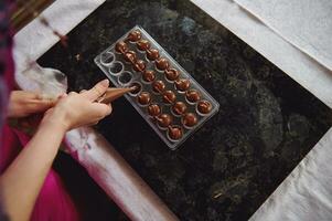 Process making handmade luxurious chocolates. Top view of a chocolatier holding a candy bag and squeezing liquid warm chocolate mass into candy molds photo