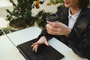 Focus on the hand holding takeaway paper mug, cardboard cup with hot drink, of a pretty woman sitting at table of outside cafe, enjoying distant remote online work, typing text on laptop keyboard photo