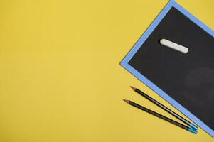 Flat lay composition with a chalk on a clean blank blackboard with copy space and two color pencils, isolated on yellow background with space for text photo