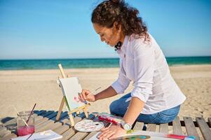 Side portrait of a beautiful female painter drawing on canvas in open air on the sea background photo