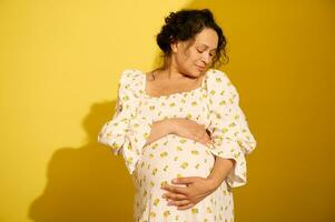 Happy gravid pregnant woman gently stroking and hugging her big belly, isolated over yellow studio background photo