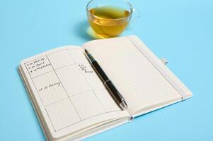 Still life of an open agenda with schedule, ink pen and transparent glass cup of tea lie on blue surface. Color background with copy space . Time management, deadline, schedule concept photo