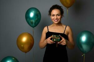 Elegant African woman in evening attire holds Christmas gift wrapped in shiny green paper and golden ribbon, cutely smiles with toothy smile looking at camera, against air balloons and gray background photo