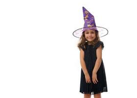 Portrait of pretty little girl wearing a wizard hat and dressed in stylish carnival dress, looking at camera posing with crossed arms against white background. Halloween concept with copy space photo