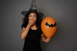 Scared woman dressed in Halloween witch costume, covering her mouth in fear and horror, looking at camera, posing with colorful balloons with felt-cut black bat against gray background with copy space photo