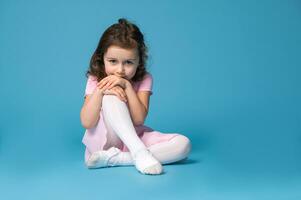 Adorable child ballerina in pink dress looking at the camera, sitting on blue background and partially covering her mouth with her hands photo
