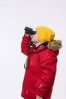 Side portrait of school boy in yellow woolen hat and bright red down jacket, looking through binoculars, isolated on white background, copy space for advertisement. Winter travel and adventure concept photo