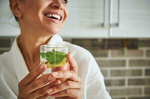 Focus on a glass with water, mint and slice of lemon in the hands of blurred woman smiling with beautiful toothy smile photo