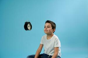 Schoolboy looking at a flying alarm clock while sitting on blue background. The concept of fleeting time. Blue background with copy space. photo
