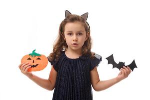 Isolated portrait on white background with copy space of adorable little girl, 4 years old kid, showing to the camera homemade felt-cut bat and pumpkin in her hands, symbol of the Halloween party photo