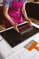 Confectioner in pink apron holding chocolate molds full of liquid heated chocolate mass and putting it on a marble surface. Making chocolates for celebrating World Chocolate Day photo