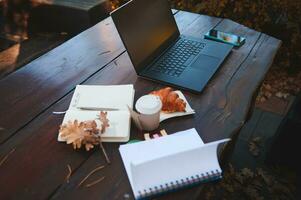 Soft focus on a laptop with copy space on blank black monitor screen near smartphone and out of focus croissant with hot coffee in takeaway paper cup and open copybook and notepad on a wooden table photo