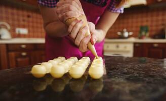 Closeup of chef in pink apron squeezing melted white chocolate with sweet creamy liquid from confectionery bag into harvested candy molds for preparing chocolate truffles photo