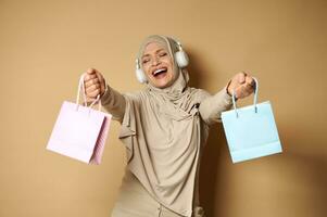 contento musulmán mujer en hijab con auriculares escuchando a música y bailando con de colores papel pantalones en su manos disfrutando el próximo religioso vacaciones. eid Mubarak dijo foto