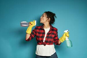 Attractive and playful young Latin American woman, a housewife, holding a cleaning spray in her hands like a gun and blowing off gunpowder as if after a shot photo