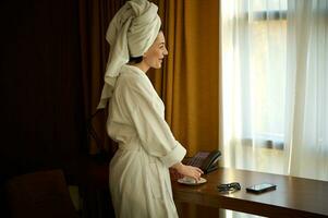 hermosa confidente negocio mujer en bata de baño y cabeza envuelto en toalla en pie por el ventana en hotel habitación y disfrutando el aroma de café en el Mañana antes de reuniones durante su negocio viaje foto