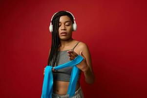 Young female athlete, African American sportswoman wearing wireless headphones wrapping a blue elastic band on her hand while standing on a red background with a copy of space photo