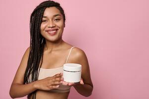 Beautiful African woman with stylish dreadlocks smiles looking at camera, holding a container with moisturizer in her hand photo