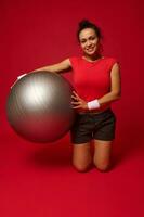 Charming African American athletic woman exercises with fit ball, smiles looking at camera isolated over red background with copy ad space photo
