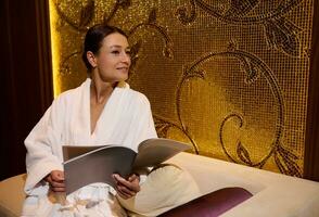Beautiful relaxed dark-haired European middle aged woman dressed in white terry bathrobe, holding a magazine and looking aside resting on a couch in a lounge room of a luxurious wellness spa complex photo