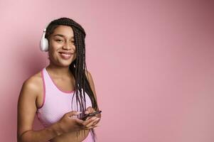 Close-up portrait of a young African woman wearing wireless headphones, using her smartphone while posing against pink background with copy space photo