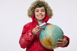 Around the world travel and winter tourism concept. Adorable elementary aged boy in warm bright red down jacket with hood holding an airplane toy model and globe, imitating the flight around the Earth photo